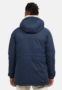 Mörkblå isolerad jacka med huva, med fleecekrage, ribbade ärmslut och en logotyp-patch på nedre ryggen. Slät textur.