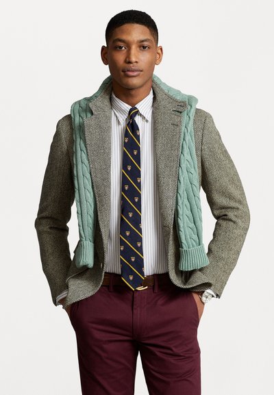 Sacou tip houndstooth cu un pulover din tricot cu cabluri de culoare verde deschis, aruncat peste o cămașă cu dungi, cravată bleumarin și pantaloni bordo.