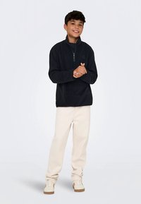 Fekete gyapjú pulóver félcipzáras gallérral, krémszínű, laza szabású nadrággal és világos lábbelivel párosítva. Simaságú felület, alkalmi design.