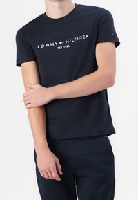 Persona vistiendo una camiseta negra de Tommy Hilfiger con el logotipo y el texto "EST. 1985", de pie con un brazo cruzado sobre el otro.