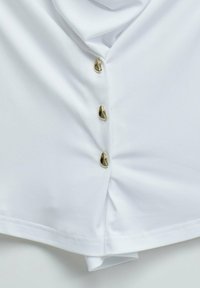 White fabric with three small irregular gold buttons fastened vertically on a gathered seam.