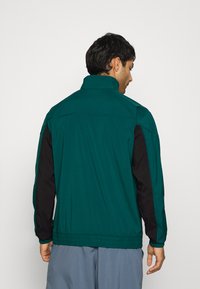 Chaqueta verde azulado con mangas negras, que presenta un collar alto, dobladillo elasticado y textura suave. Diseñada para un ajuste relajado.