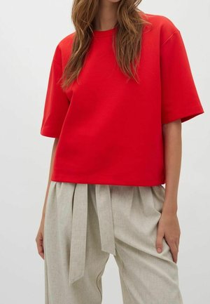 Femme portant un haut ample rouge vif à manches courtes et un pantalon beige clair taille haute avec une ceinture nouée.