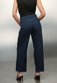 Vaqueros de mezclilla azul oscuro de talle alto con pierna recta, que cuentan con bolsillos traseros y lazos laterales en el bajo. Textura suave, diseño casual.