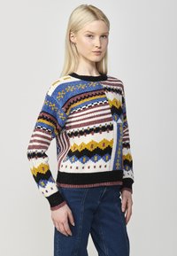 Pull en tricot coloré avec des motifs géométriques en bleu, jaune et bordeaux, doté d'un col et de poignets côtelés noirs.