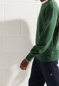 Osoba oblečená v zeleném tričku s dlouhým rukávem, tmavě modrých kalhotách s malým logem a stříbrným náramkem na zápěstí, stojí před bílou cihlovou zdí.