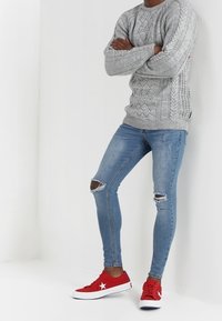 Grauer Strickpullover mit Zopfmuster, zerrissene blaue Skinny-Jeans und rote Sneakers mit weißen Akzenten. Lässiges Outfit, das Textur und Farbkontrast betont.
