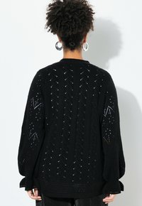 Černý pletený cardigan s odhalovaným vzorem, který má prodloužené rukávy a strukturovaný ribbing lem. Lehký a volný střih.