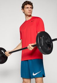Rød atletisk t-shirt med en sort logo, parret med blå shorts med en hvid Nike swoosh. Modellen holder en sort vægtstang.