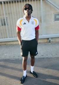 Camisa da Alemanha da Adidas com detalhes em vermelho e amarelo, calções pretos, meias brancas e loafers pretos. Conjunto atlético casual com um design em contraste.