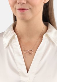 Roséguld oändlighetshalsband med ett hjärtanhänge, med en polerad yta och en delikat kedja, buren över en vit skjorta med krage.