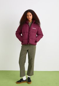 Burgundy puffer jacket with quilted design and logo, paired with olive green corduroy pants and black sneakers with brown soles.