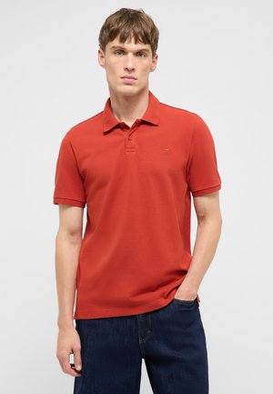 Junger Mann trägt ein rotes kurzärmeliges Poloshirt und dunkelblaue Jeans, steht mit einer Hand in der Tasche vor einem einfarbigen hellen Hintergrund.