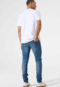 Witte t-shirt met korte mouwen, gecombineerd met blauwe slim-fit jeans met achterzakken en minimalistische stiksels, gedragen met lichte sneakers.