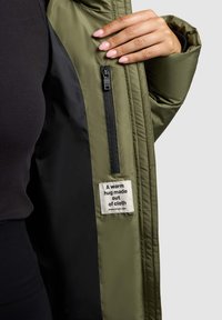 Olivgrüne Steppjacke mit einer Reißverschlusstasche und einem Stoffetikett mit der Aufschrift "Eine warme Umarmung aus Stoff", ausgestattet mit einer glatten, glänzenden Oberfläche.