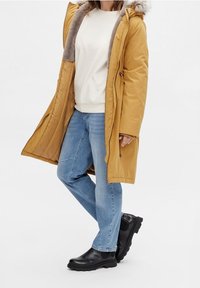 Hořčicově žlutá zateplená bunda s kožíškovou kapucí, zkombinovaná s lehkým svetrém, modrými džíny a černými kotníkovými botami. Hladký, izolovaný materiál.