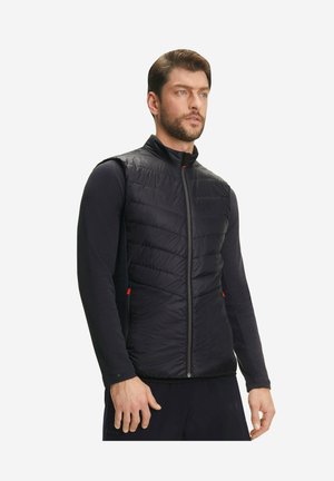 Uomo che indossa un gilet nero trapuntato senza maniche sopra una camicia a maniche lunghe, in piedi contro uno sfondo bianco uniforme, rivolto leggermente verso destra.