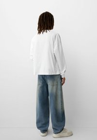 Bijela košulja dugih rukava opuštenog kroja u kombinaciji s prevelikim svijetloplavim trapericama, koje imaju gradijentni pranje i široke nogavice. Bijele tenisice.