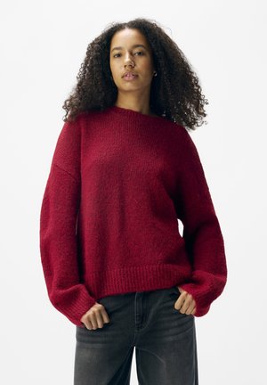 Red knit sweater with a textured, loose-fit design; wide sleeves and ribbed hem; paired with dark gray jeans, against a plain white background.