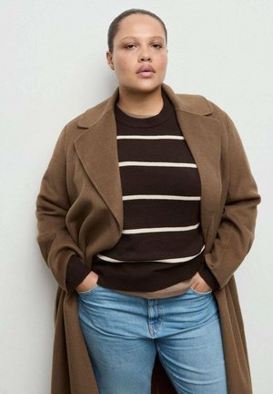 Długi brązowy płaszcz na brązowym swetrze w paski oraz jasnoniebieskich dżinsach. Płaszcz ma szerokie klapy, a sweter ma fakturę dzianiny.