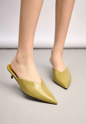 Pieds portant des escarpins en cuir jaune-vert à bout pointu, à enfiler, avec de petits talons kitten sur une surface grise.