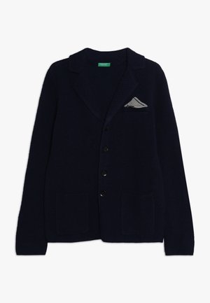 Granatowy sweter z dzianiny z kołnierzem typu szal, pięcioma czarnymi guzikami, dwoma kieszeniami z przodu oraz małym szarym detalem w postaci chusteczki w kieszeni.