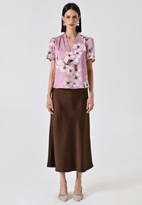 Blusa floreale rosa con maniche a palloncino corte, abbinata a una gonna marrone fino al ginocchio e scarpe beige con punta arrotondata. Tessuti morbidi.