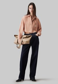Camicia button-up a maniche corte di colore pesca con taschino sul petto, abbinata a jeans a gamba larga blu scuro e una borsa strutturata beige tenuta da un manico.