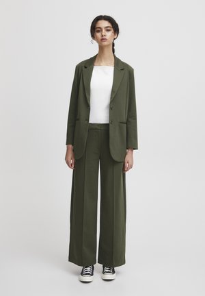 Olijfgroene blazer en wijde broek in zachte stof, gecombineerd met een getailleerd wit topje en zwarte sneakers met zilveren accenten.