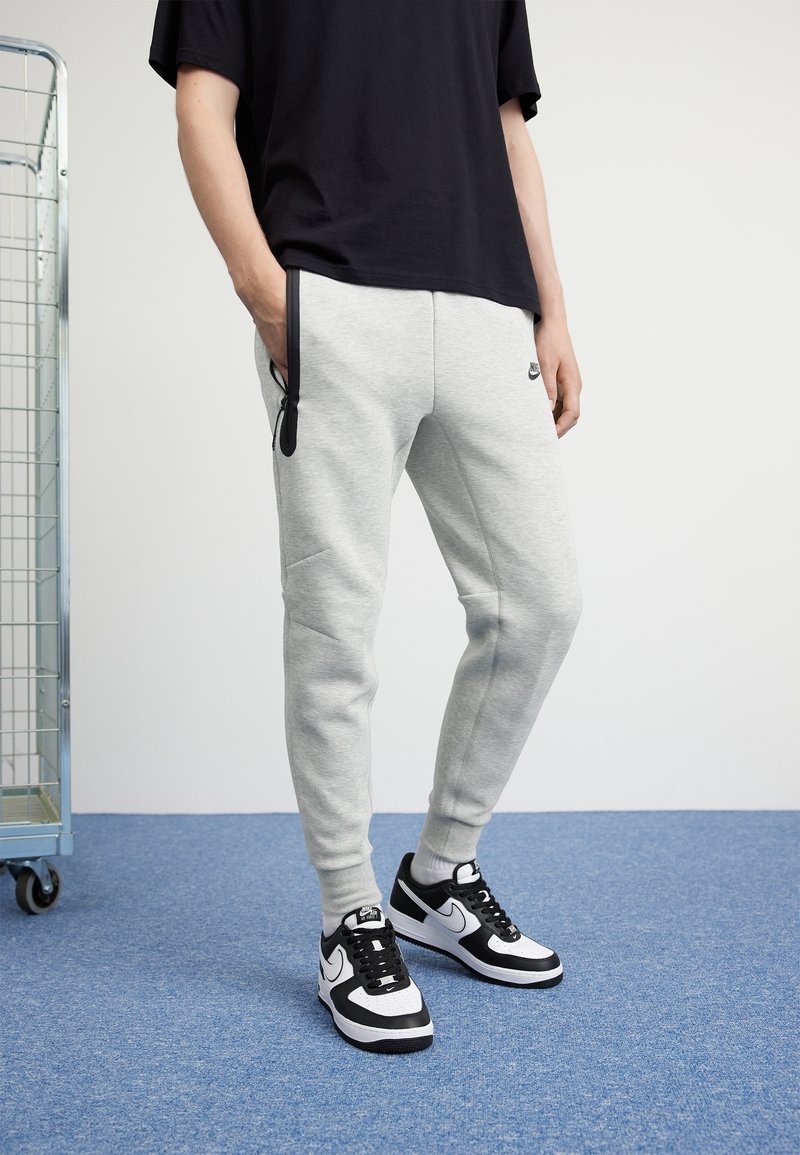 Grå atletiska sweatpants med en smidig design, som har svarta detaljer och en sidoficka, i kombination med svart-vita sneakers.