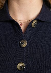 Primo piano di un cardigan lavorato a maglia scuro con tre bottoni marmorizzati e una collana dorata con un ciondolo a croce di pietra rossa.