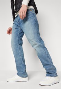 Jeans de mezclilla de color azul claro con corte recto. Llevados con unas zapatillas blancas que tienen acentos perforados y un logo de color en los jeans.