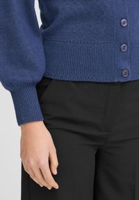 Sötétkék kardigán puha kötött anyagból, bordázott mandzsettával és szegéllyel, öt lila gombbal az elején, fekete nadrággal párosítva.
