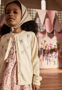 Cream cardigan with blue ribbon embroidery, textured fabric, and buttons, paired with a floral dress featuring ruffles. Background with fabric decorations.