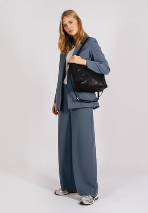 Costume bleu avec un pantalon large et un blazer, accompagné d'un sac en cuir noir. Des baskets argentées complètent le look, sur un fond uni.