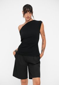 Top nero a spalla unica realizzato in tessuto morbido, caratterizzato da un design drappeggiato e una silhouette aderente, abbinato a pantaloni corti neri larghi.