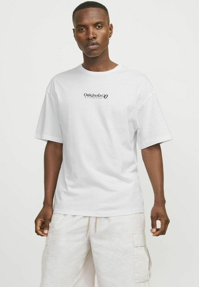Camiseta de algodón blanca con cuello redondo, mangas cortas y un logotipo negro de "Originals Studio" impreso en el pecho. Diseño de corte clásico.