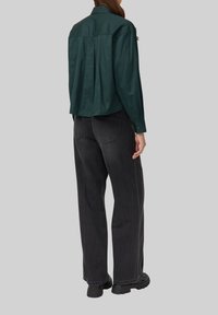 Chemise verte à manches longues avec un détail de pli au dos, associée à un jean large noir et des bottes noires épaisses, vue de dos.