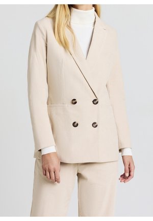 Donna che indossa un blazer doppiopetto in velluto a coste beige e pantaloni abbinati, con dolcevita bianco sotto, in piedi davanti a uno sfondo semplice.