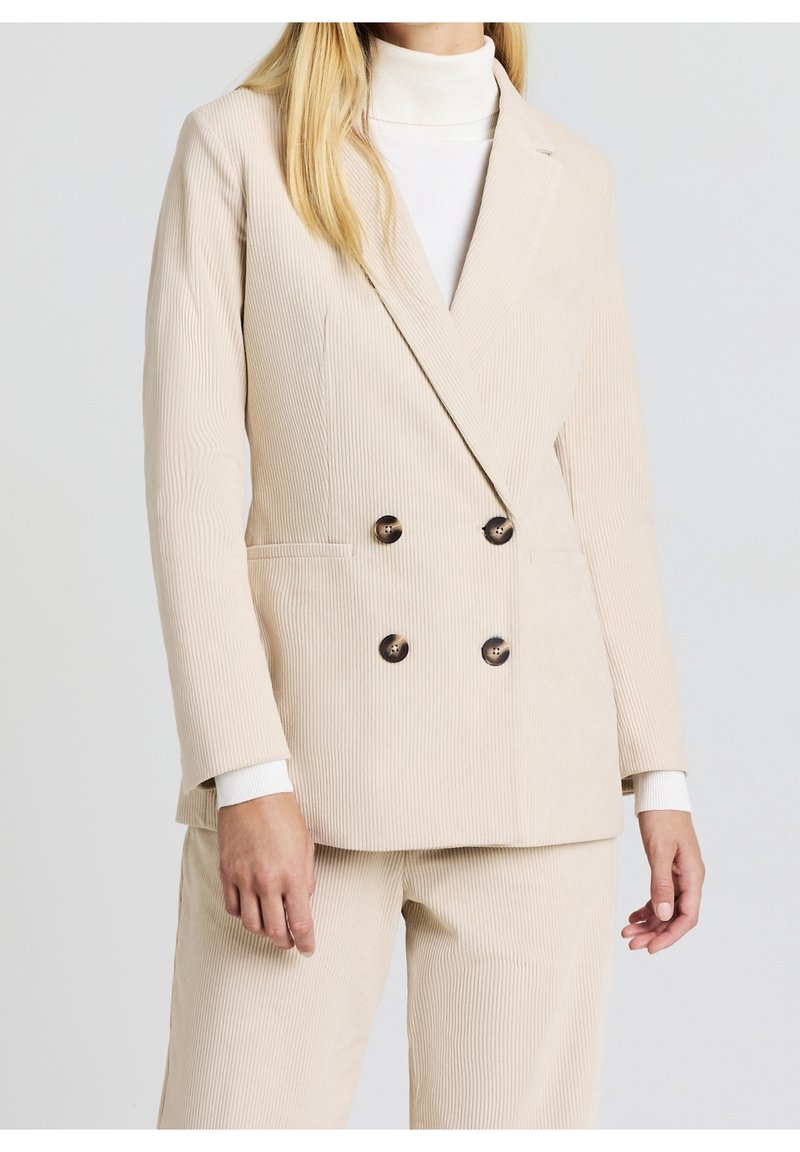 Femme portant un blazer en velours côtelé beige à double boutonnage et un pantalon assorti, avec un col roulé blanc en dessous, debout devant un fond uni.