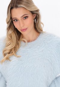 Blonde Frau mit lockigem Haar, die silberne Creolen und einen hellblauen, flauschigen Pullover mit kleinen Kristallen trägt, schaut nach vorne.