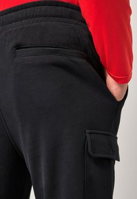 Osoba nosící černé cargo kalhoty s boční kapsou a zadní průhmatovou kapsou, ruka v kapse, oblečená v červeném dlouhém rukávu.