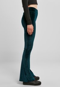 Tirkizne plišane hlače na zvono s visokim strukom, glatke teksture i uskog kroja, u kombinaciji s crnim cipelama.