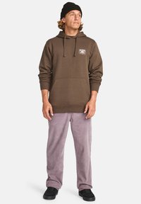 Homme portant un sweat à capuche marron avec poche avant, un bonnet noir, un pantalon violet clair et des chaussures noires, se tenant et regardant légèrement vers la droite sur fond blanc.