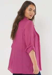 Blusa ligera de color rosa con un ajuste relajado, mangas enrolladas y una tela texturizada. Presenta una pestaña abotonada en la manga.