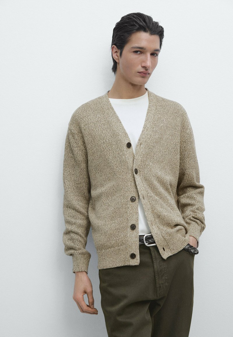 Young man wearing a beige buttoned cardigan over a white shirt, dark green pants, and a braided black belt, standing against a white wall.