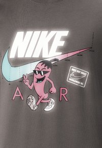 Šedé tričko s výrazným bílým logem "NIKE", růžovou kreslenou postavičkou a tyrkysovým swoosh. Doplňuje ho růžový a černý text.