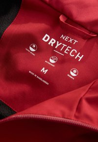 Röd jacka med etikett som visar "Next Drytech," storlek medium, tillverkad i Bangladesh, med ikoner för tejpade sömmar, vattentålighet och värmeegenskaper.