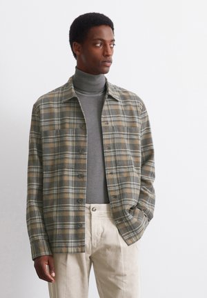 Grahamuster in Grau mit braunem und weißem Karomuster, Knopfleiste vorne, kombiniert mit einem grauen Rollkragenpullover und hellen Cordhosen.