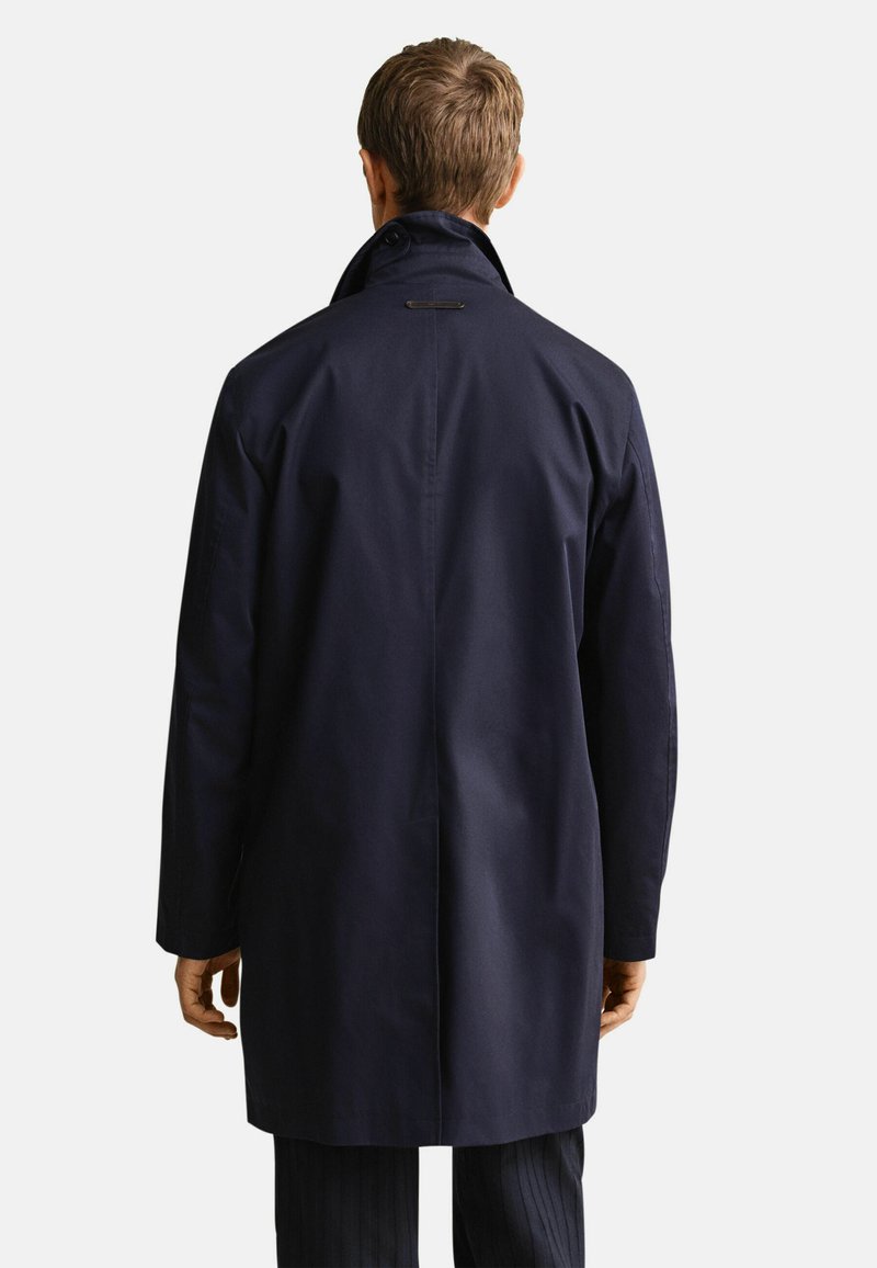 Ein Mann, von hinten gesehen, trägt einen mittel-langen dunkelblauen Mantel mit hochgestelltem Kragen über dunklen Hosen vor einem einfarbigen hellen Hintergrund.